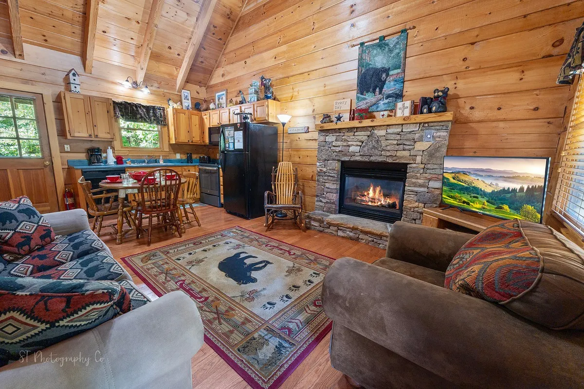 This image captures the heart of the cabin's living space, showcasing a cozy fireplace, a dining table ready for family gatherings, a charming rug with an animal motif, and a clear view of the adjacent kitchen area. The warm wooden tones and inviting atmosphere highlight the seamless flow between relaxation, dining, and meal preparation.
