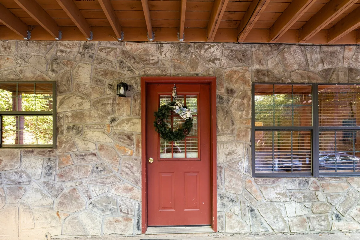 An inviting exterior view of the cabin featuring a bold red door adorned with a festive wreath set in a rustic stone wall. The entryway hints at a welcoming home base after a day of outdoor adventure.