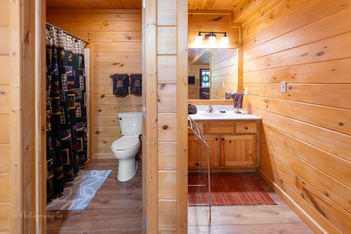 A warmly lit bathroom with wooden walls, a vanity mirror, toilet, and shower curtain, exuding a cozy, private feel. The space appears inviting and convenient, perfect for refreshing after a day of play.