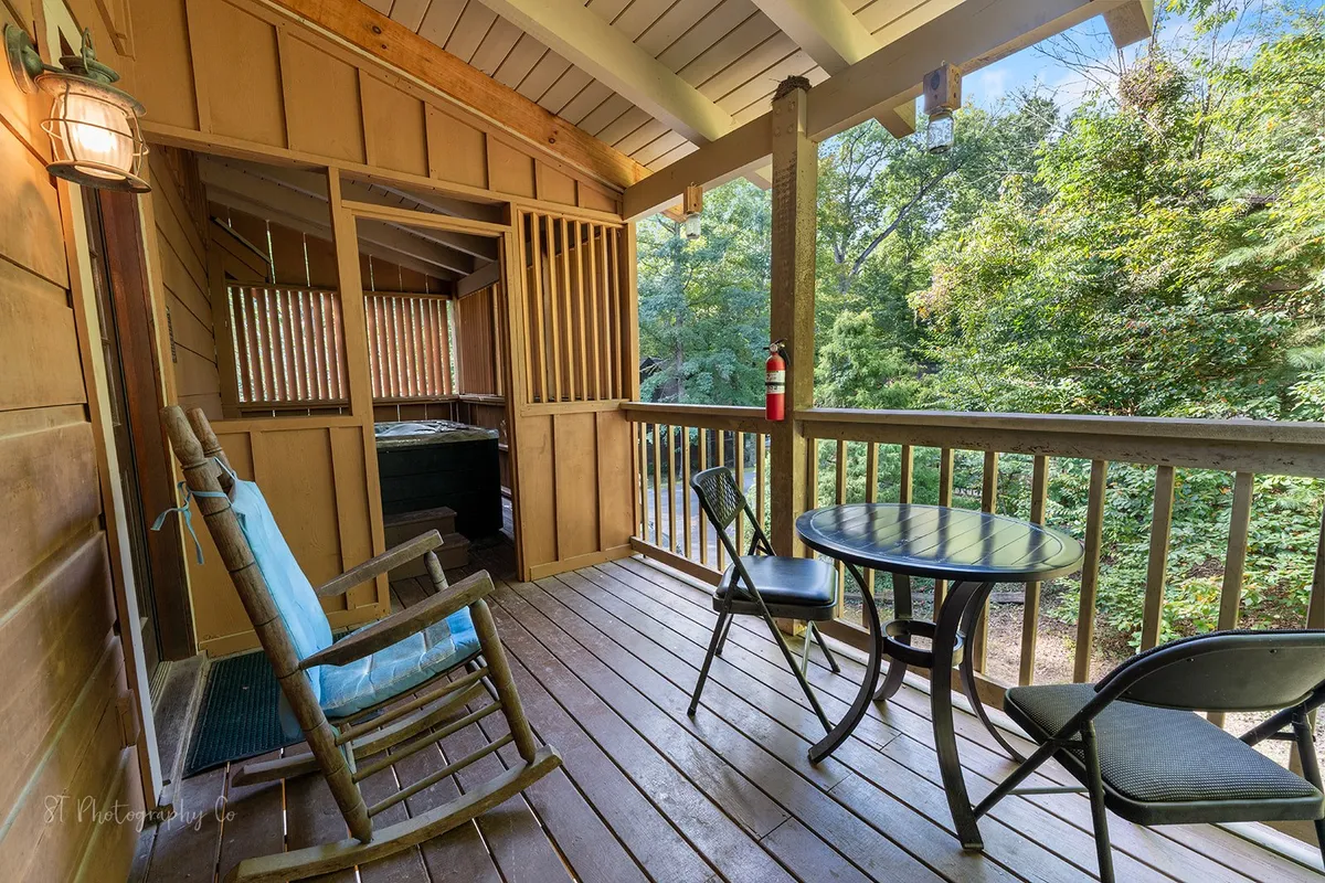 This image shows a spacious covered deck featuring a relaxing hot tub, a comfortable rocking chair, a table with chairs, and lush greenery surrounding the area. The atmosphere is calm and inviting, perfect for unwinding and enjoying the outdoors.