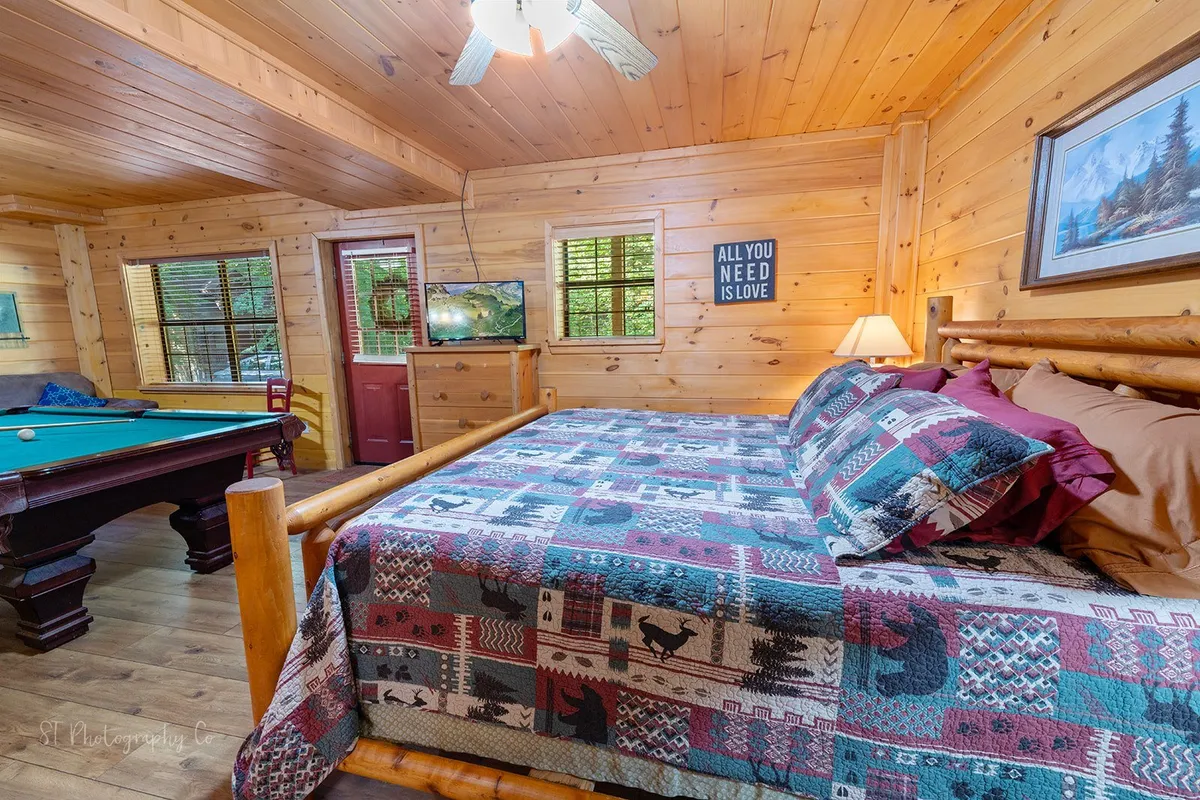 This warm, friendly scene spotlights a bed with a patterned quilt and a pool table nearby, set beneath a wooden ceiling with framed artwork and a window showing greenery outside. The setup suggests an intimate, multi-functional space ideal for both play and rest.