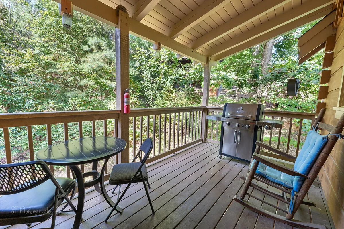 A cozy outdoor deck with a grill, rocking chair, table with chairs, and lush greenery all around. The setup is ideal for both relaxation and socializing, with a pet-friendly vibe suggested by the spacious, open layout.