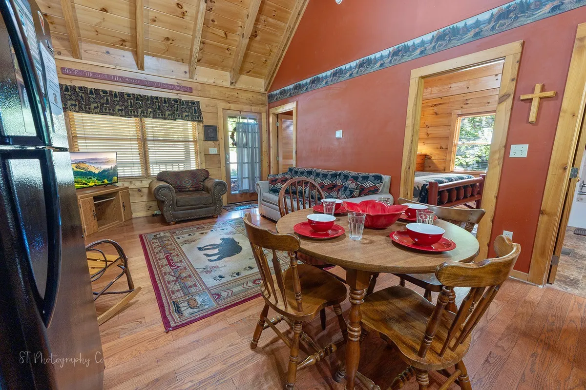This image features a comfortable sofa area with patterned cushions, a dining table set with red plates, a large TV displaying a scenic image, and timber beams and walls that give the space a rustic yet inviting feel. The arrangement illustrates how the living room, dining, and kitchen interact as a cohesive gathering hub.