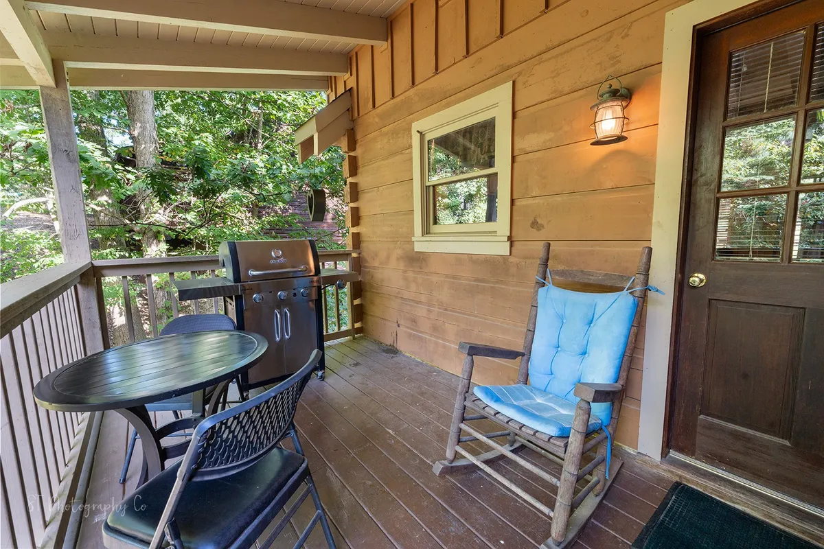 A spacious outdoor area featuring a rocking chair, a table with chairs, a grill, and natural wooden siding. The porch is surrounded by greenery, offering a tranquil setting perfect for group cookouts or relaxing after exploring local attractions.