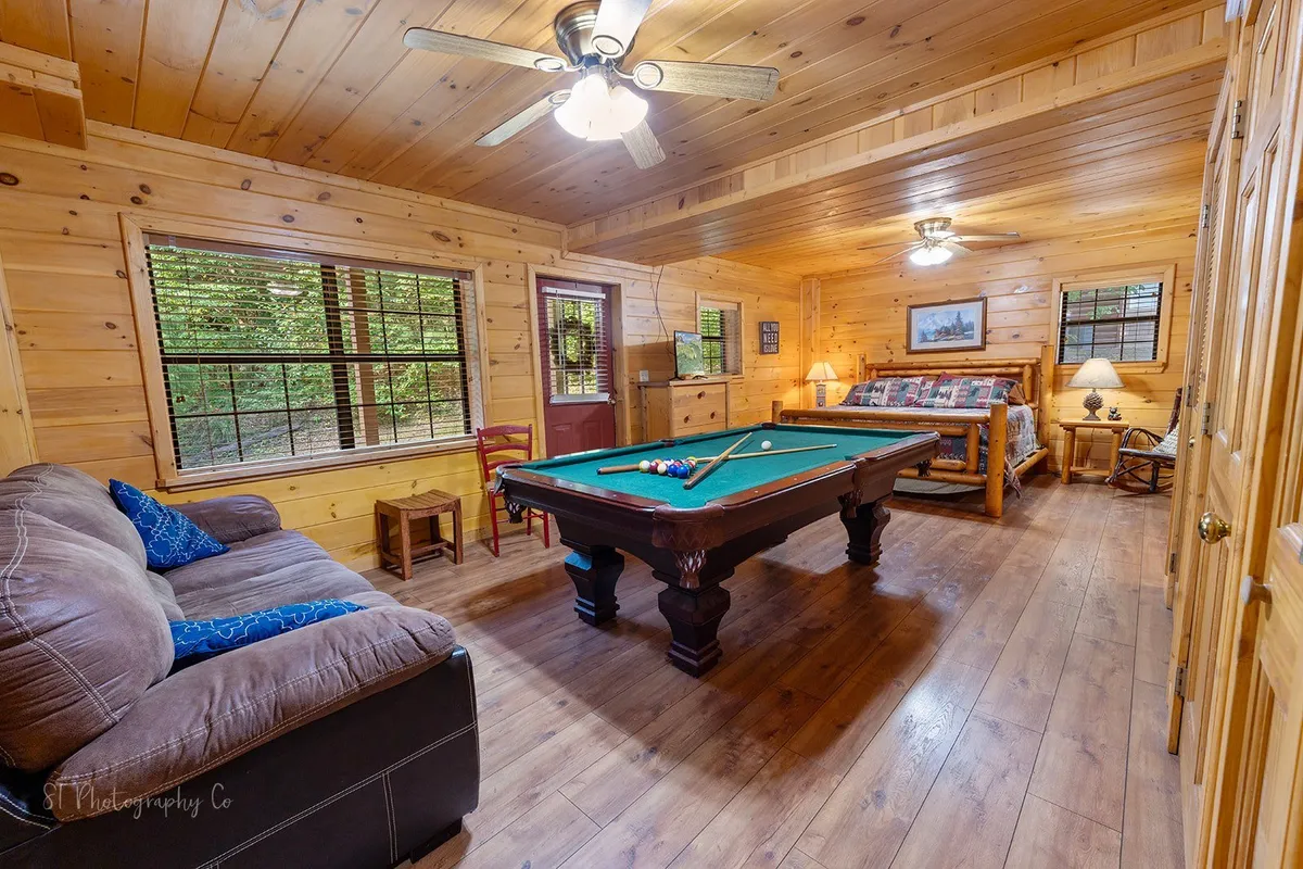 This image captures a spacious interior featuring a pool table at the center, a comfortable sofa, a king-sized bed, and large windows letting in natural light. The arrangement clearly merges gaming and lounging areas within a single room, echoing the suite's versatile and inviting atmosphere.
