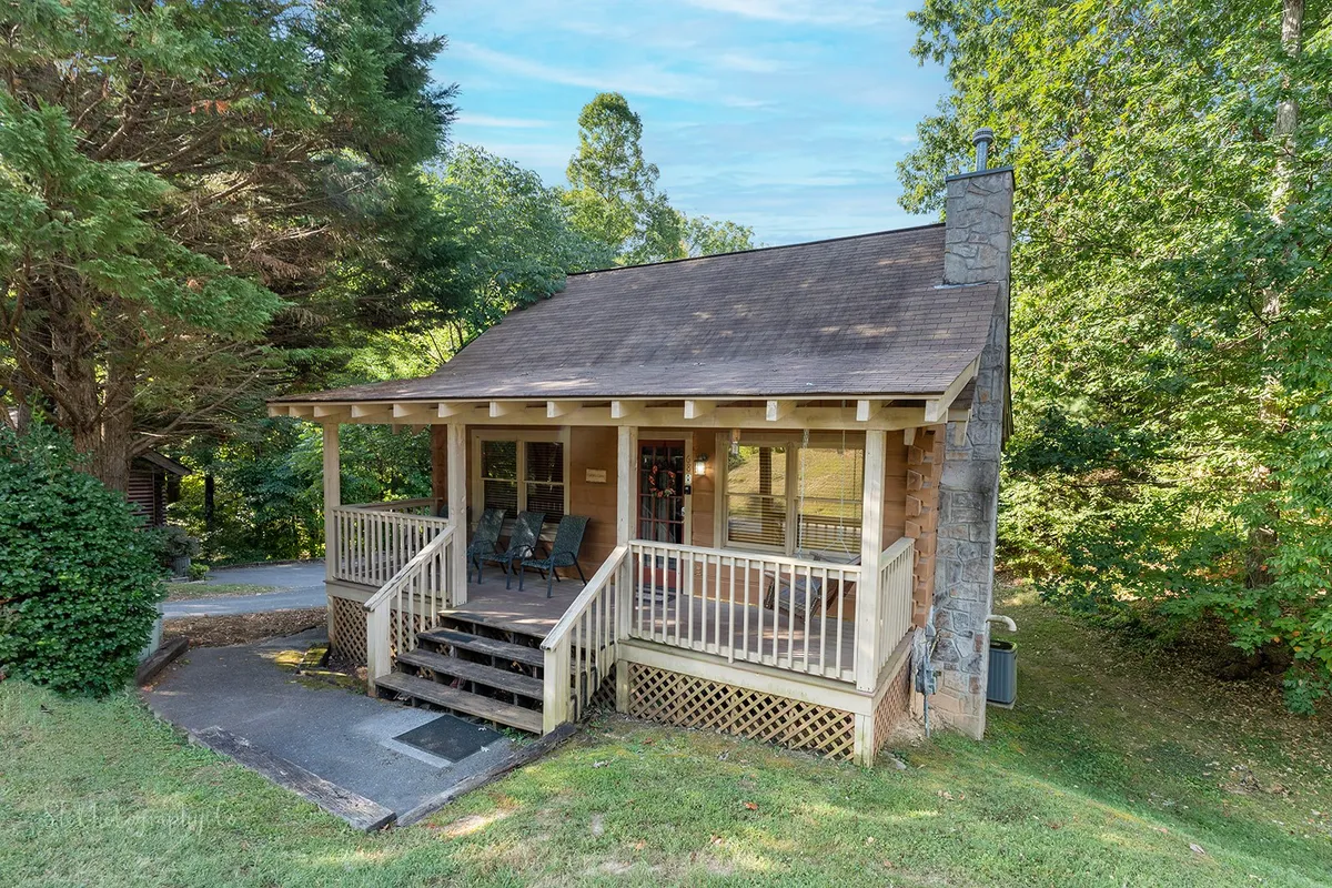 This image shows the inviting front porch of Karen's Cabin, featuring wooden steps, a sturdy stone chimney, and lush greenery surrounding the property. The porch offers comfortable seating for relaxation and socializing after a day of exploring local attractions.